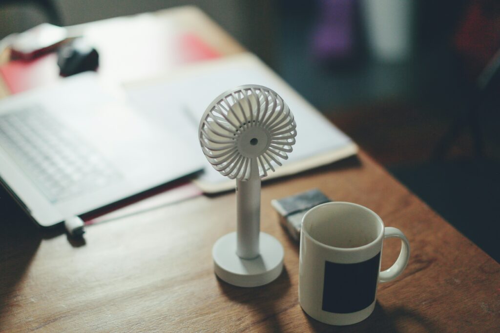 Tischventilator zum Schutz vor Hitze bei der Arbeit auf Schreibtisch mit Tasse im Vordergrund. Im Hintergrund ist ein unscharfer Laptop zu sehen
